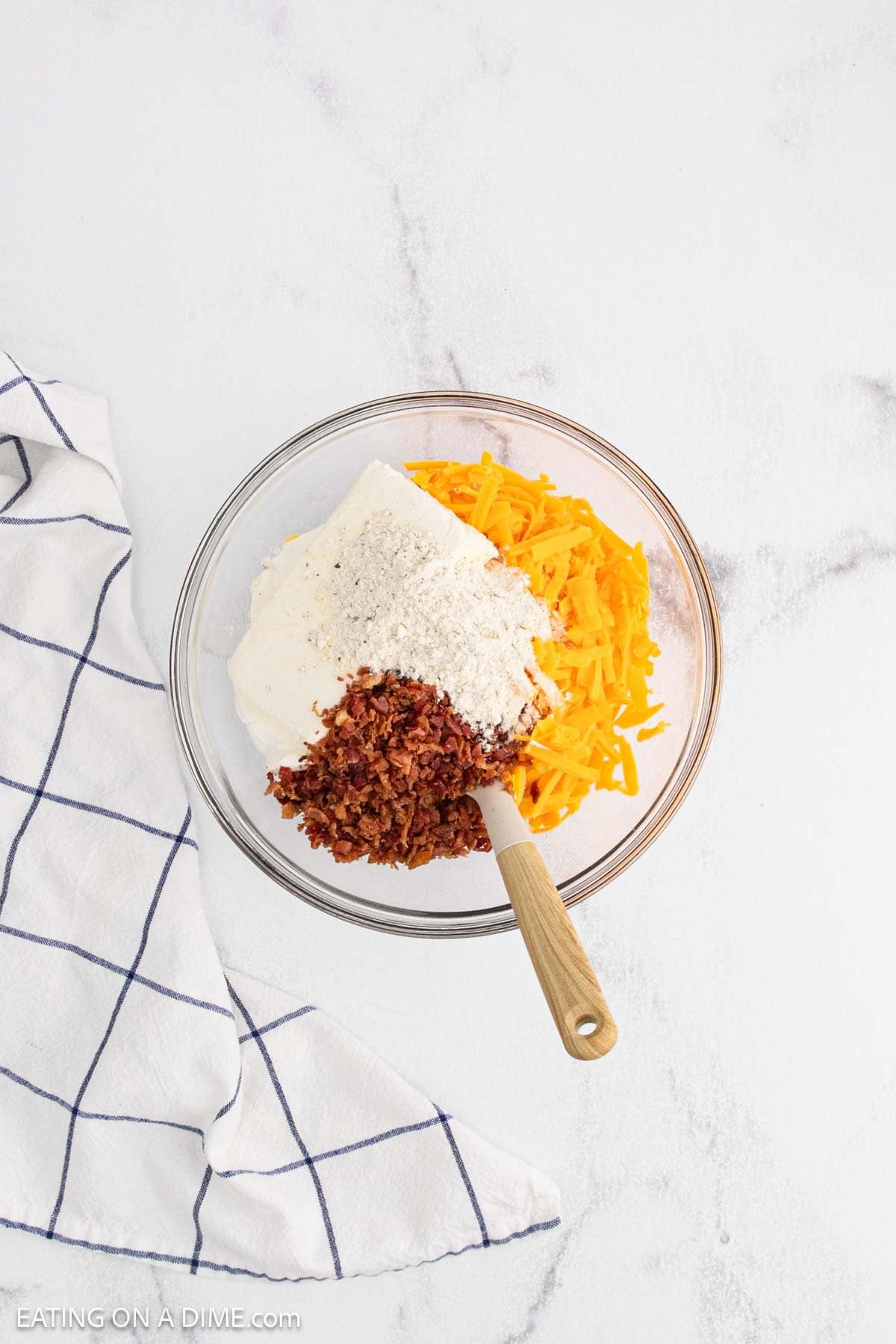 A glass mixing bowl on a white surface contains shredded cheddar cheese, crumbled bacon, cream cheese, and ranch seasoning—the classic ingredients for a delicious Crack Dip. A wooden spatula rests in the bowl. A white towel with blue lines lies nearby.