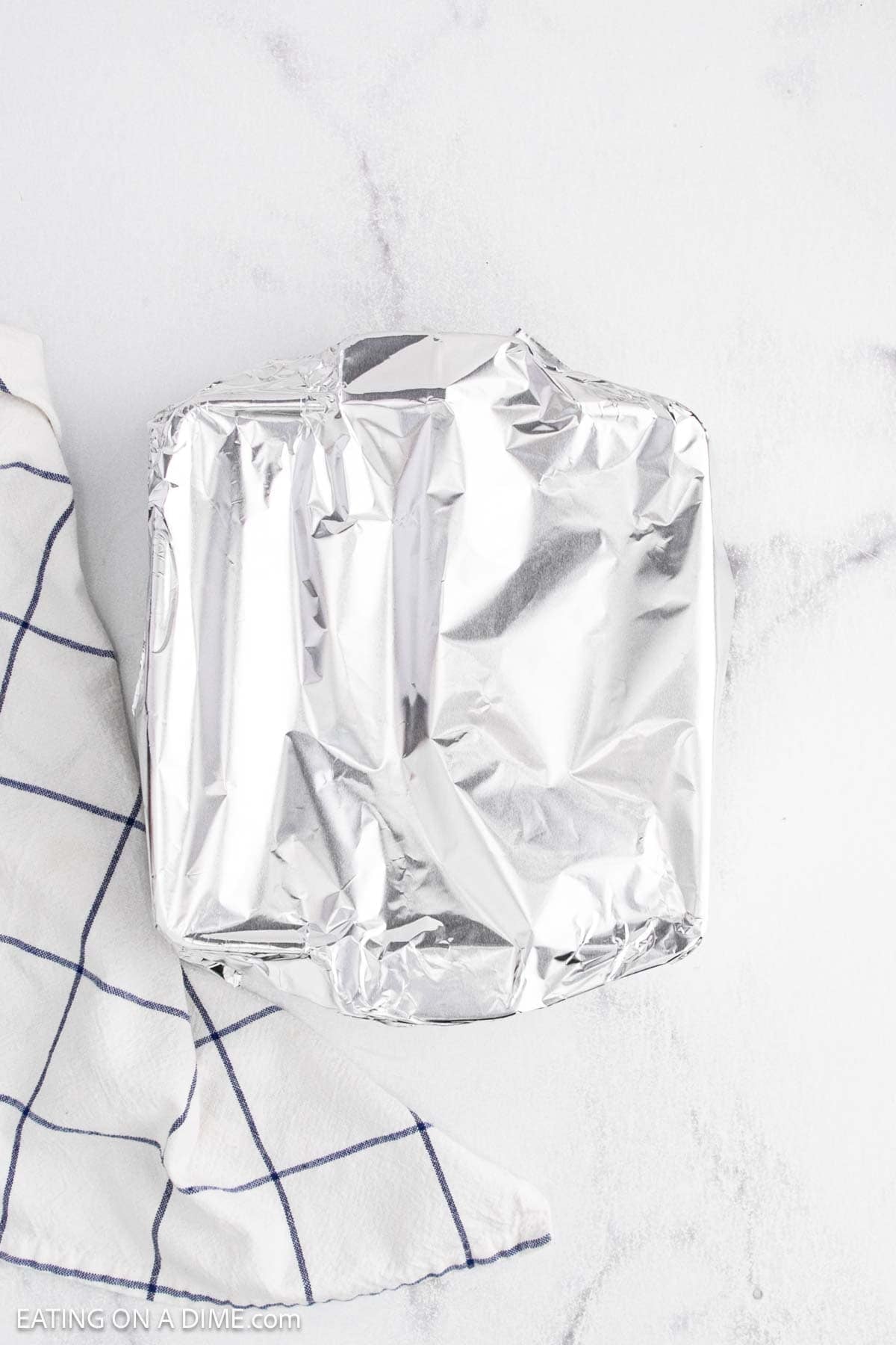 A square baking dish covered with aluminum foil sits on a white surface next to a white kitchen towel with blue grid lines, ready to keep your favorite Crack Dip warm and delicious.
