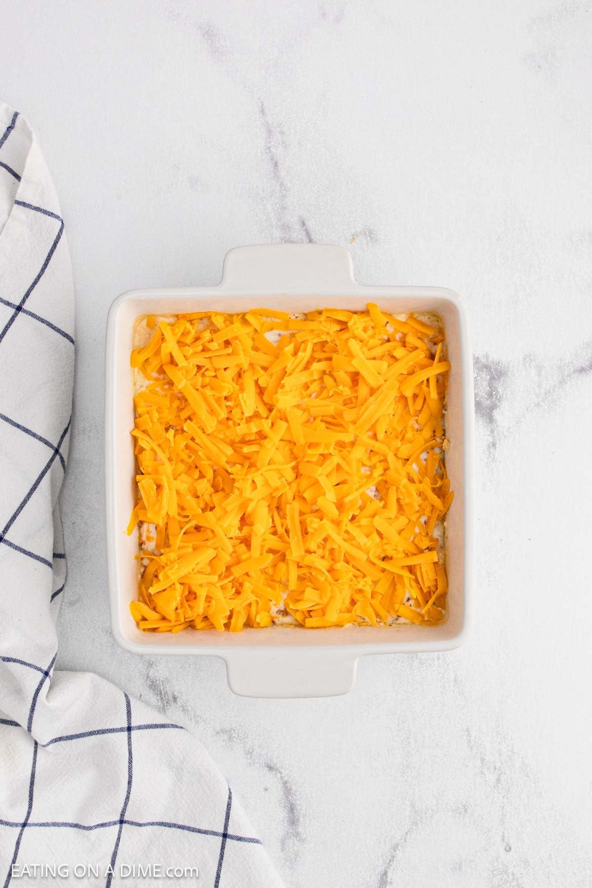 A square white baking dish filled with shredded cheddar cheese for Crack Dip sits on a white countertop next to a white kitchen towel with a blue grid pattern.