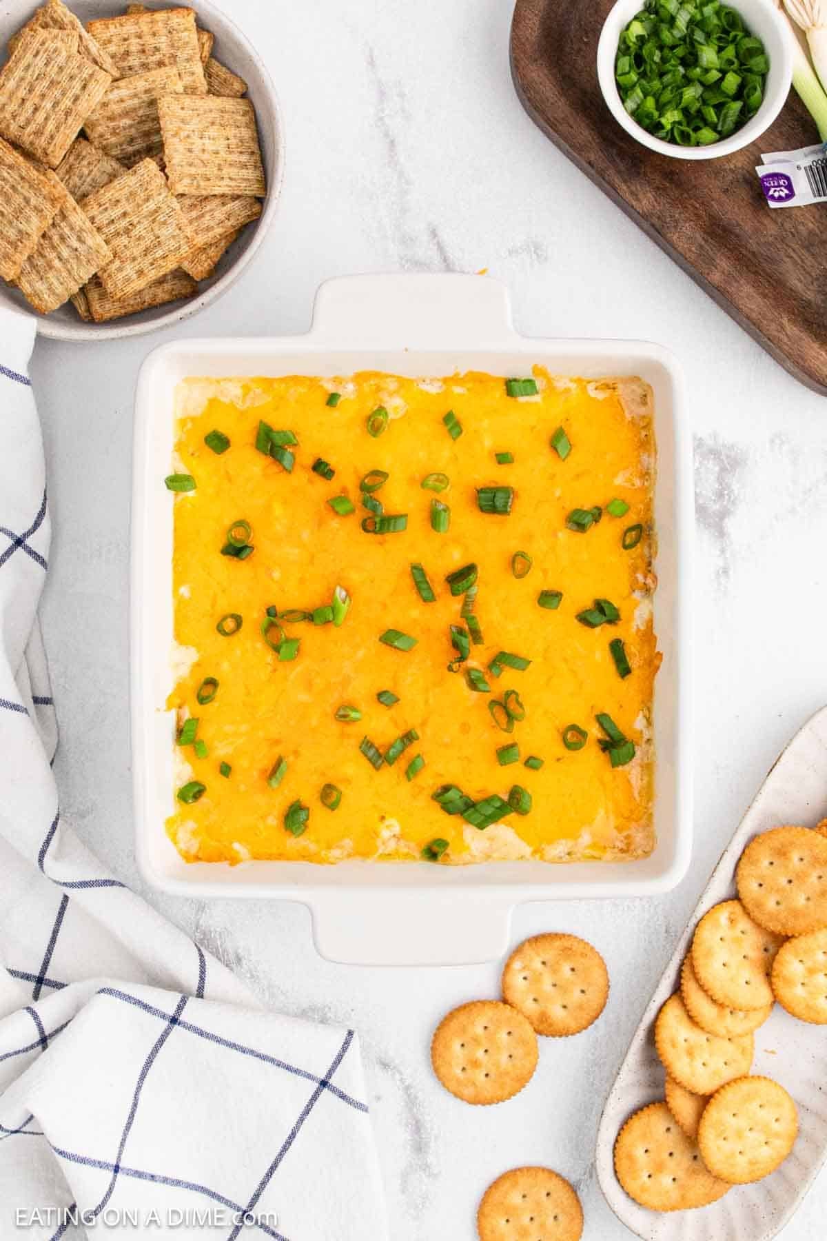 A square white baking dish filled with melted cheesy Crack Dip, topped with chopped green onions. Surrounding the dish are round crackers, woven wheat crackers, a napkin, and a small board with more chopped green onions.