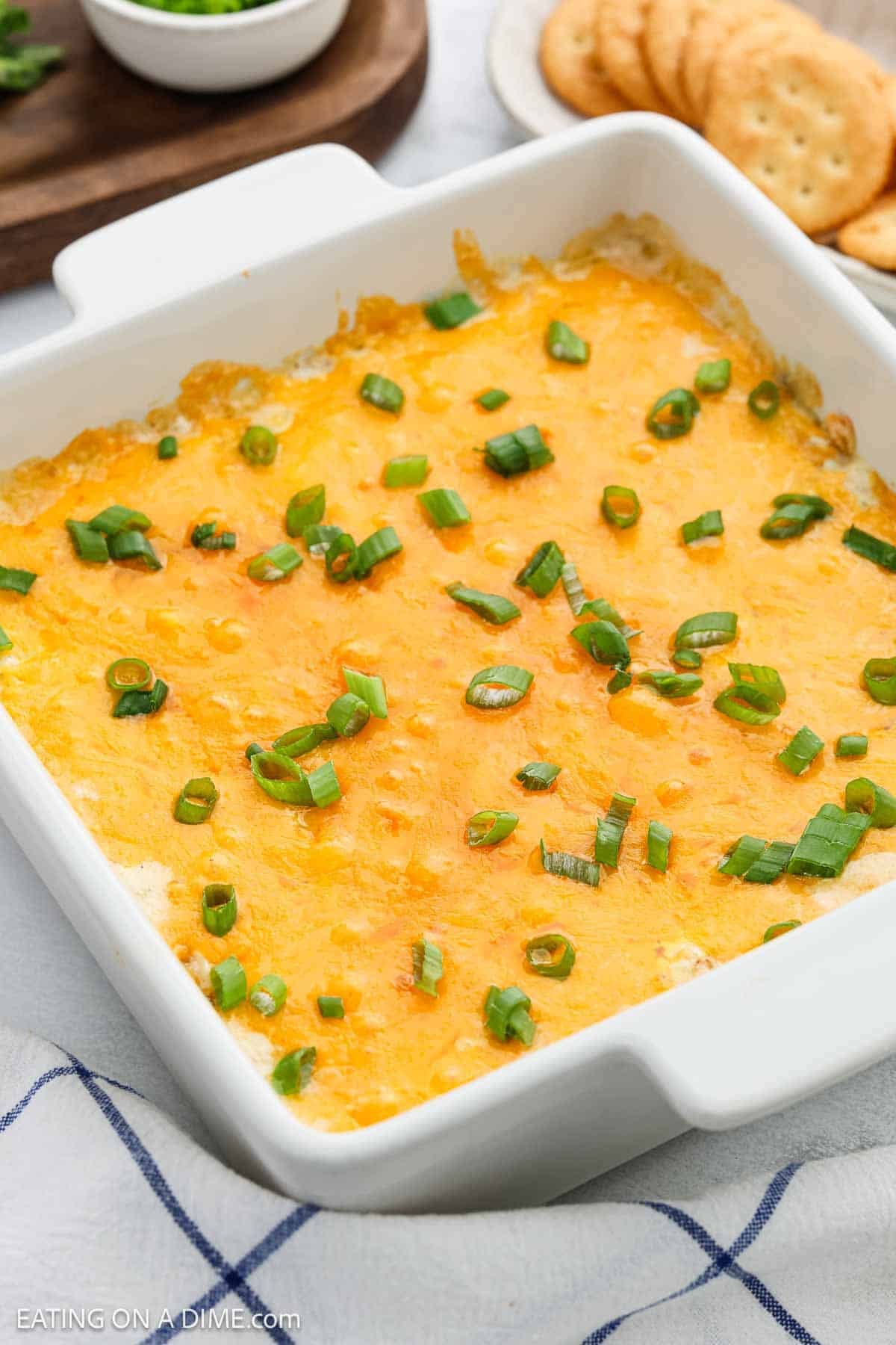 A white baking dish filled with Crack Dip, topped with melted cheddar and chopped green onions, is surrounded by crackers and a cloth napkin.