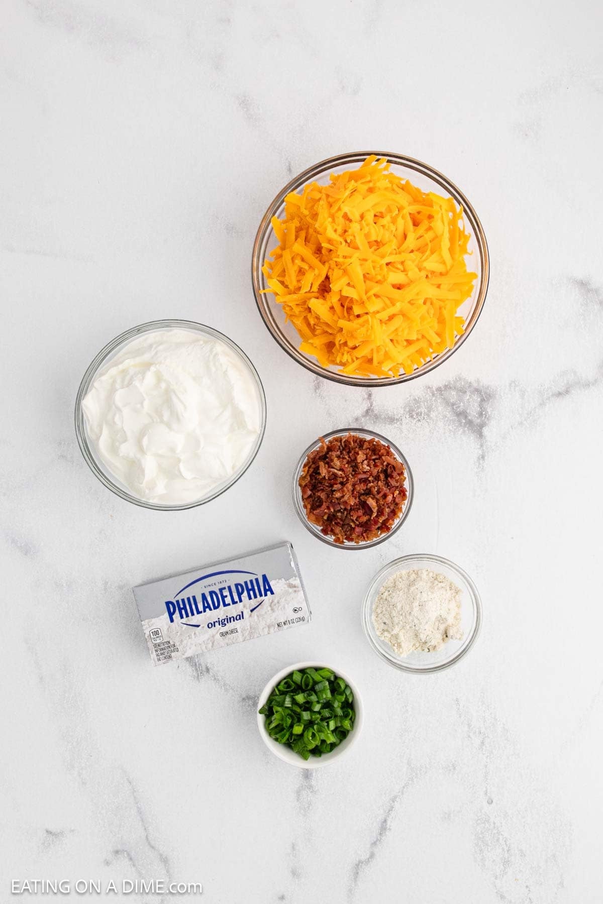Five bowls and containers on a white surface hold shredded cheddar cheese, sour cream, bacon bits, chopped green onions, a block of cream cheese, and a small bowl of white seasoning—perfect ingredients for making delicious Crack Dip.
