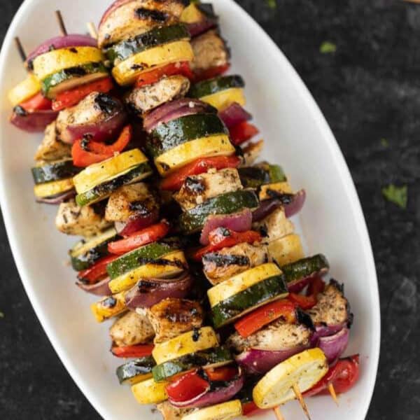 Grilled chicken kabob and vegetable skewers are arranged on a white oval plate, featuring slices of zucchini, yellow squash, red onion, red bell pepper, and seasoned grilled chicken chunks.