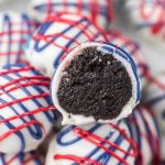 Close-up of a Patriotic Oreo Truffle with a white candy coating, adorned in red and blue drizzle. The bitten truffle reveals a dark, rich chocolate center, while more festive Patriotic Oreo Truffles sit in the background.
