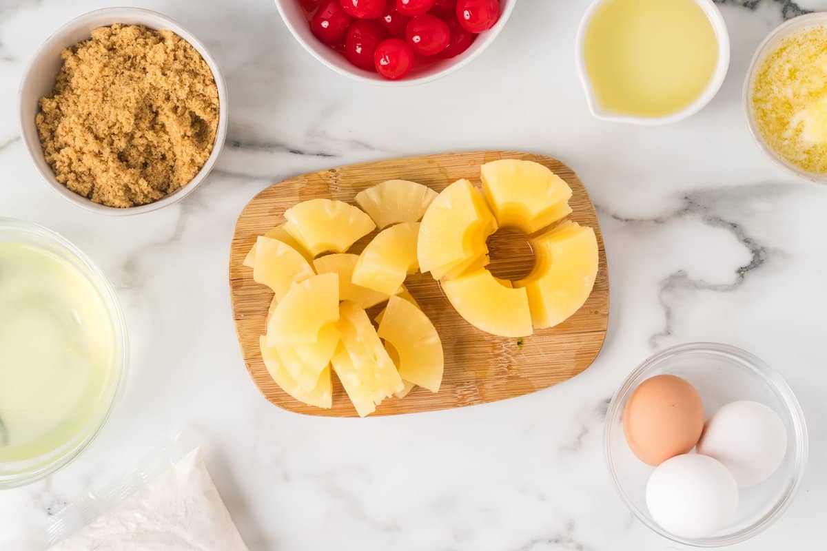 Cutting up pineapple slices on a cutting board with a bowl of eggs, brown sugar and cherries