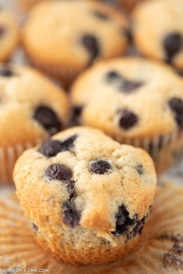 A close up of a blueberry muffin with the wrapper removed and other muffins behind it. 