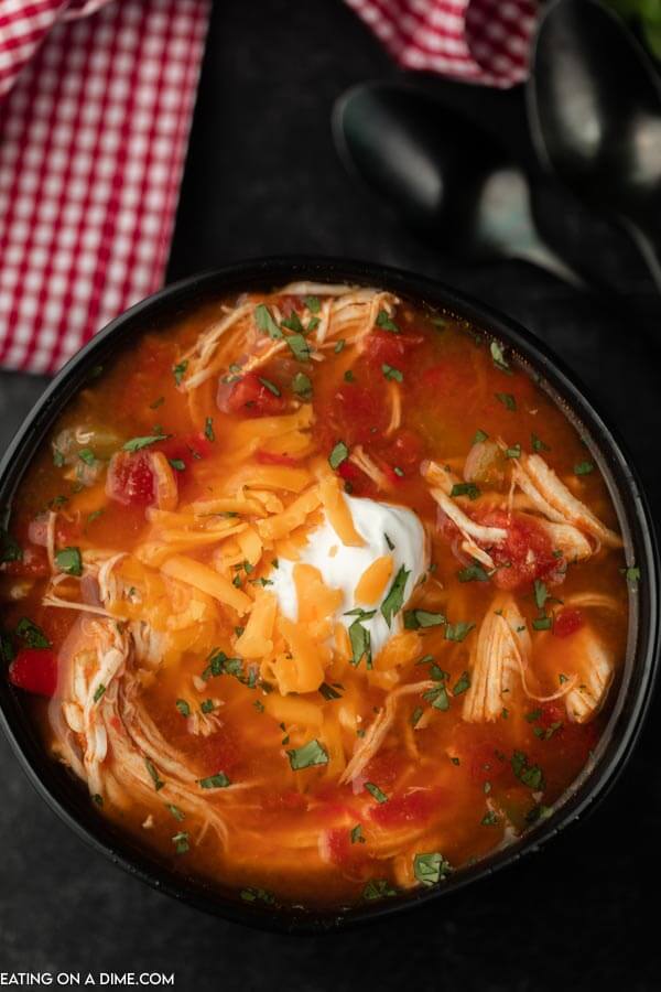 A bowl of chicken fajita soup topped with sour cream and cheddar cheese with a red and white towel next to it. 