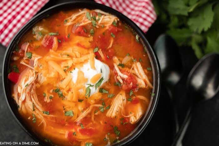 A black bowl of chicken fajita soup topped with sour cream and cheddar cheese with 2 black spoons next to it. 