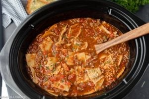 A close-up of crock pot lasagna soup recipe with noodles, ground meat, tomatoes, and herbs simmering in a black slow cooker. A wooden spoon rests in the soup, and some bread is visible in the background.