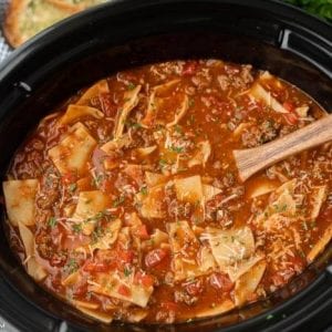 A close-up of crock pot lasagna soup recipe with noodles, ground meat, tomatoes, and herbs simmering in a black slow cooker. A wooden spoon rests in the soup, and some bread is visible in the background.