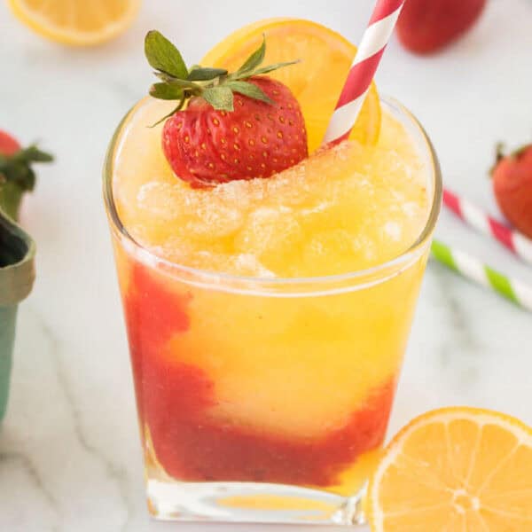 A glass of yellow-orange frozen strawberry lemonade with a red layer at the bottom, garnished with a fresh strawberry and orange slice, and served with a red-and-white striped straw. Sliced oranges and strawberries are nearby.