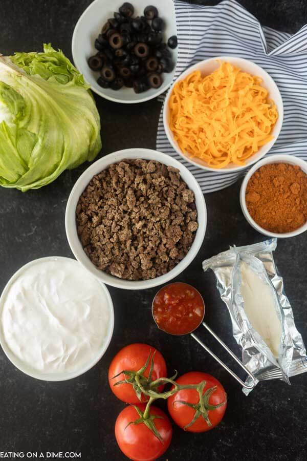 Close up image of Ingredients needed for Taco Dip - Lettuce, Hamburger Meat, Cheese, Taco Seasoning, Sour cream, Black Olives, Salsa, Tomatoes