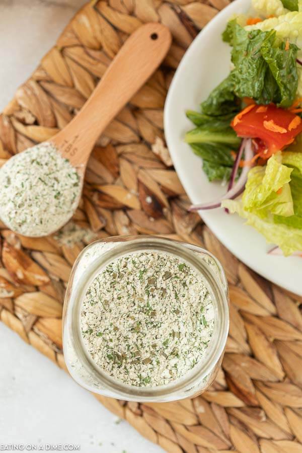 A tablespoon of the ranch dressing mix next to a jar full of the homemade ranch seasoning mix with a salad in the back ground on a woven place mat. 