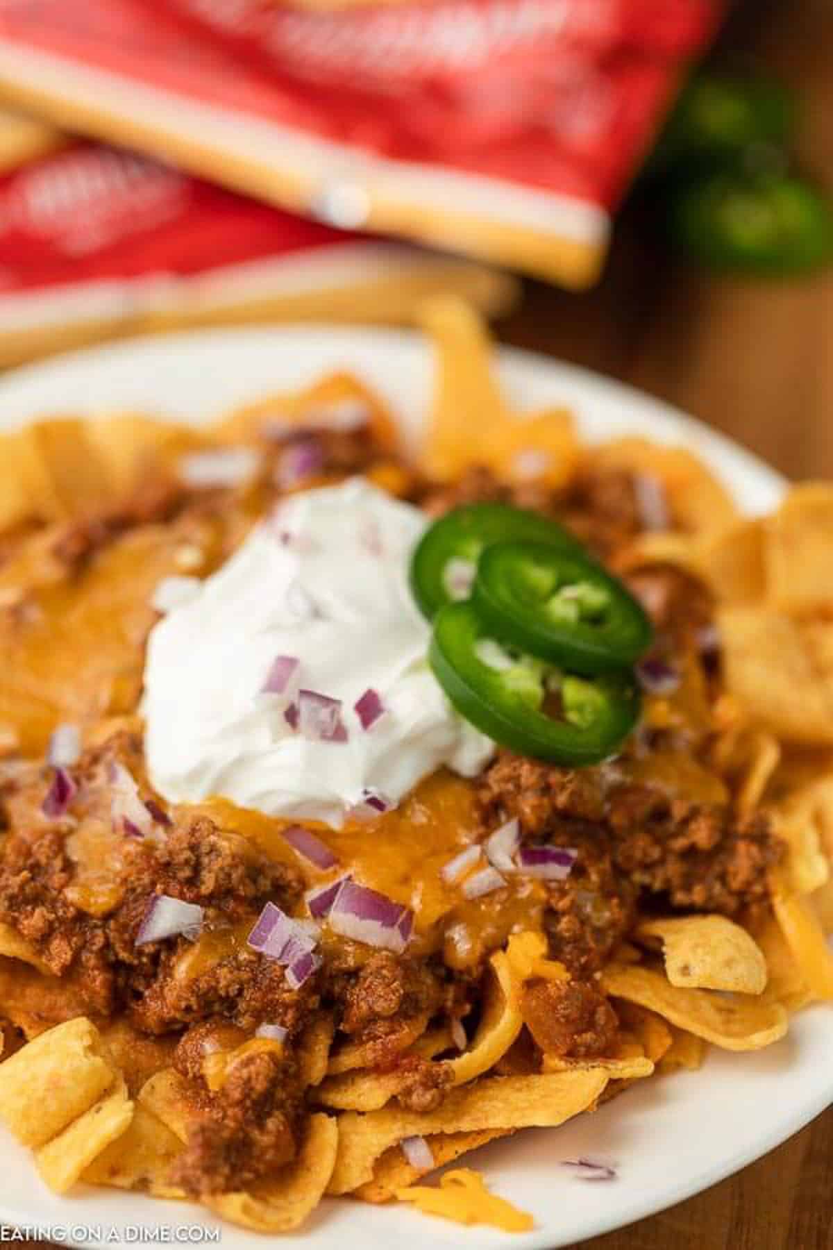 photo of frito pie on plate topped with shredded cheese, sour cream, sliced jalapeños and diced red onions 