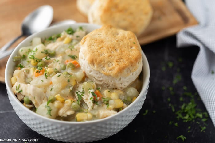 A large white bowl full of Instant Pot Chicken Pot Pie topped with a biscuit and parsley with more biscuits and spoons behind the bowl.  