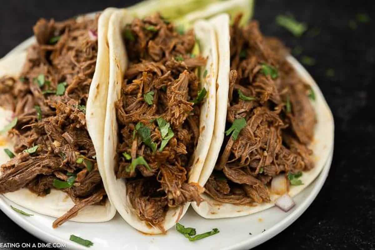 3 shredded beef tacos topped with cilantro, avocado on a white plate on a black countertop. 