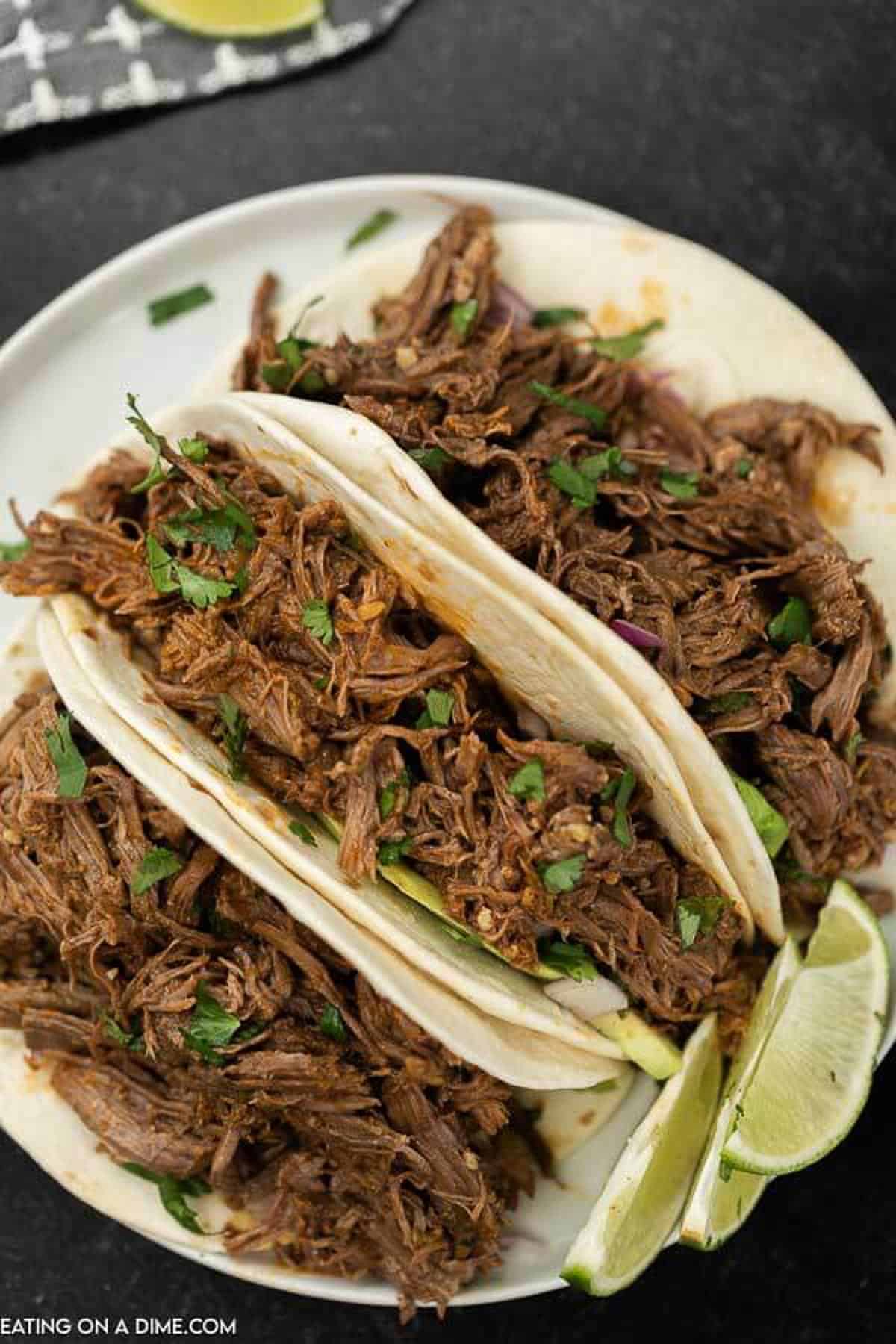 3 shredded beef tacos topped with cilantro, avocado on a white plate on a black countertop. 