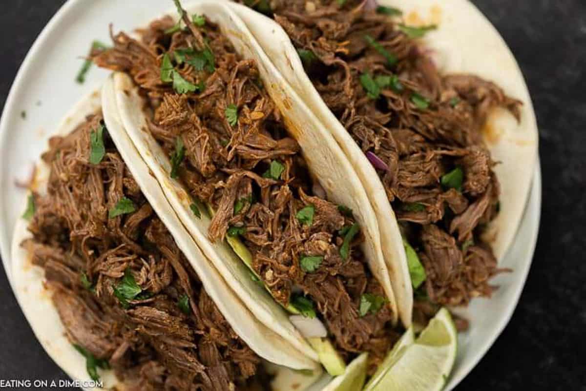 3 Shredded beef tacos on flour tortillas topped with cilantro and lime on a white plate. 