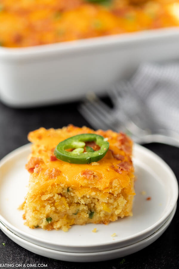A piece of the jalapeño popper corn casserole on a plate in front of the casserole in a pan.  