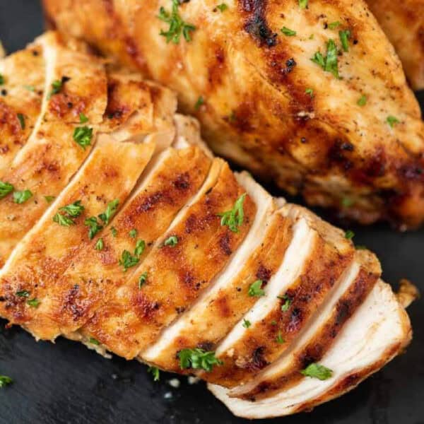 Sliced grilled chicken breast garnished with chopped parsley, showing a juicy and golden-brown exterior, perfect for adding to your favorite leftover grilled chicken recipes. Served on a dark surface.