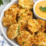 A plate of crispy, golden-brown Air Fryer Fried Pickles is arranged next to a small bowl of creamy dipping sauce, garnished with parsley.