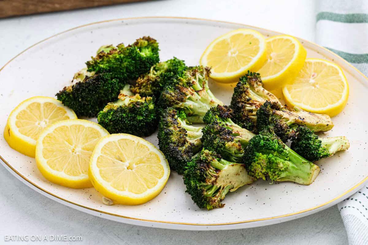 A white plate with grilled broccoli florets and several slices of fresh lemon arranged on it. The food is seasoned and displayed on a light-colored surface.