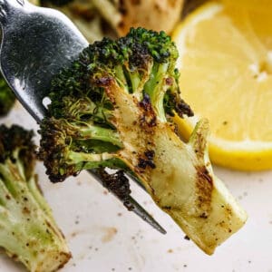 A close-up of grilled broccoli on a fork, showcasing charred grill marks, with a sliced lemon in the background.