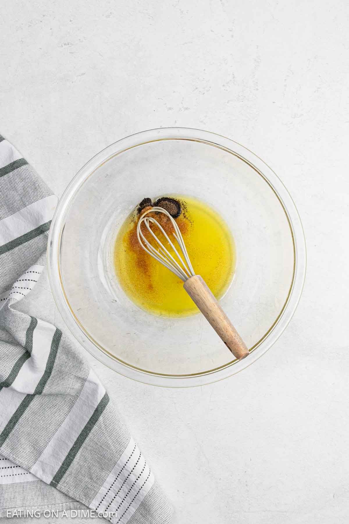 A glass mixing bowl with melted butter and spices being whisked together for grilled broccoli, placed on a white surface next to a gray and white striped kitchen towel.
