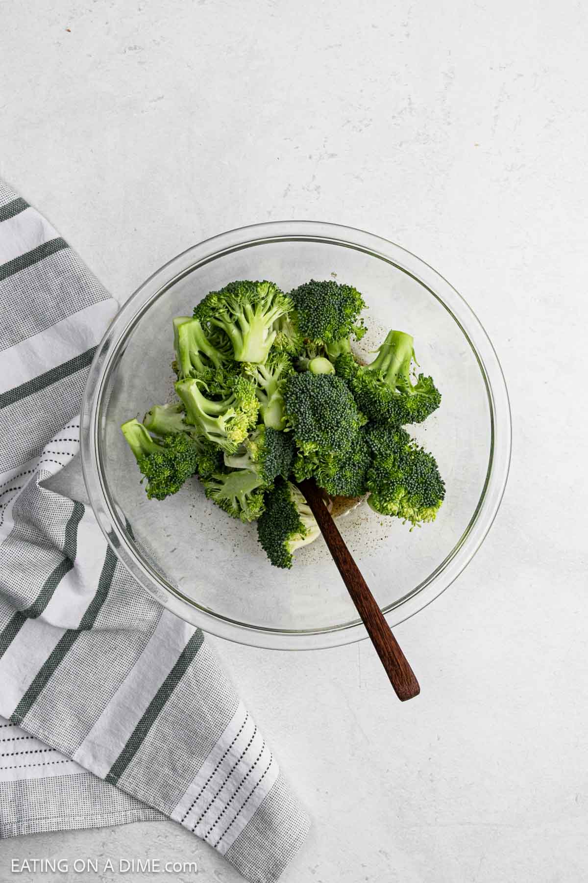 A clear glass bowl filled with fresh broccoli florets sits on a light surface, next to a striped kitchen towel. A wooden spoon is in the bowl, ready for mixingโperfect for prepping grilled broccoli.