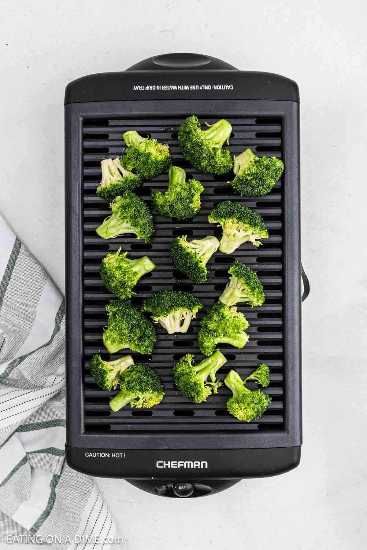 Grilled broccoli florets are arranged on a black electric grill with a striped towel beside it, set on a light-colored surface.