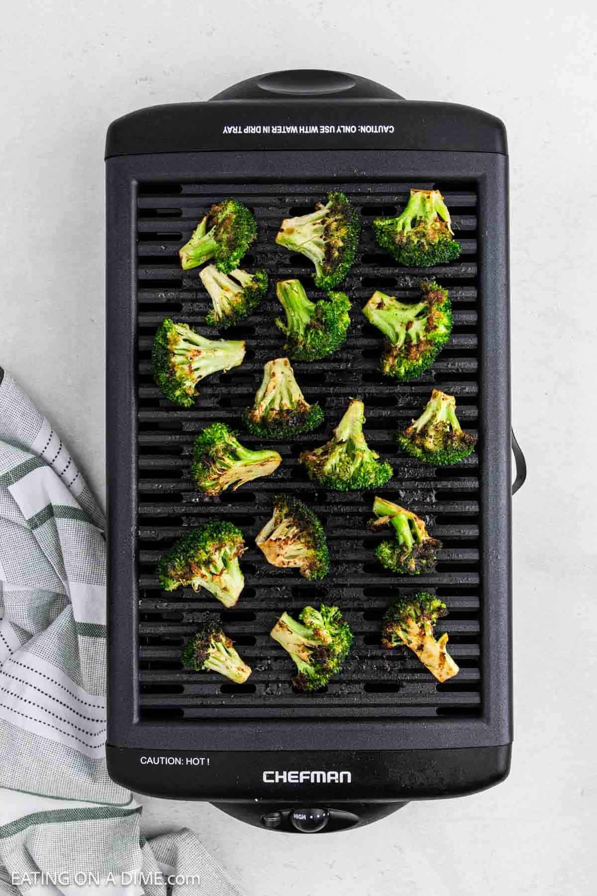 Grilled broccoli florets are spread out on a black electric Chefman grill, with a striped kitchen towel visible to the side on a light-colored countertop.