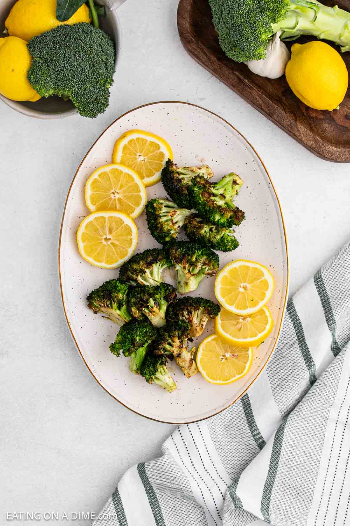Grilled broccoli florets are arranged on an oval plate, garnished with fresh lemon slices. A striped cloth napkin, whole broccoli, lemons, and garlic are seen nearby on a light countertop.