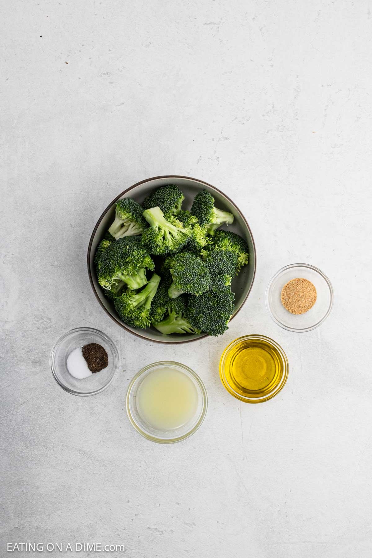 A bowl of fresh broccoli florets sits next to small bowls containing olive oil, lemon juice, seasoning, and salt and pepperโperfect ingredients for making grilled broccoliโarranged on a light-colored surface.
