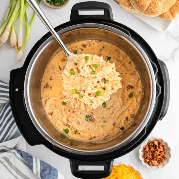 Overhead view of creamy shredded chicken in an Instant Pot, garnished with green onions. Surrounding the pot are ingredients like shredded cheese, bacon bits, green onions, and sandwich buns—perfect for this instant pot crack chicken recipe.