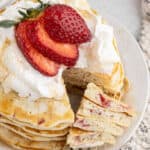 A stack of fluffy strawberry pancakes topped with whipped cream and sliced strawberries, with a wedge cut out and placed at the side, showing the airy texture inside.