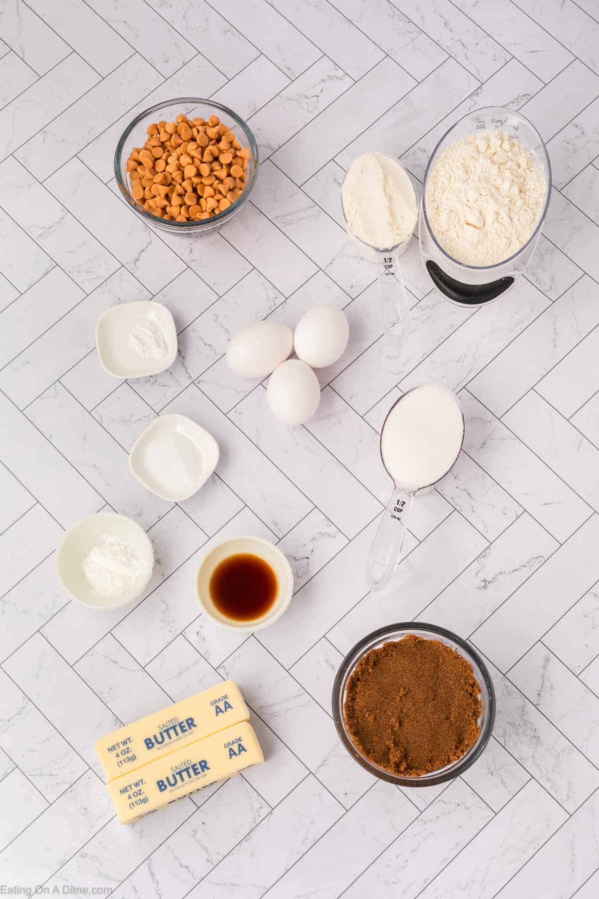 A variety of baking ingredients arranged on a white tiled surface, perfect for making butterscotch brownies, including eggs, butter sticks, brown sugar, granulated sugar, flour, butterscotch chips, vanilla extract, baking powder, and baking soda.