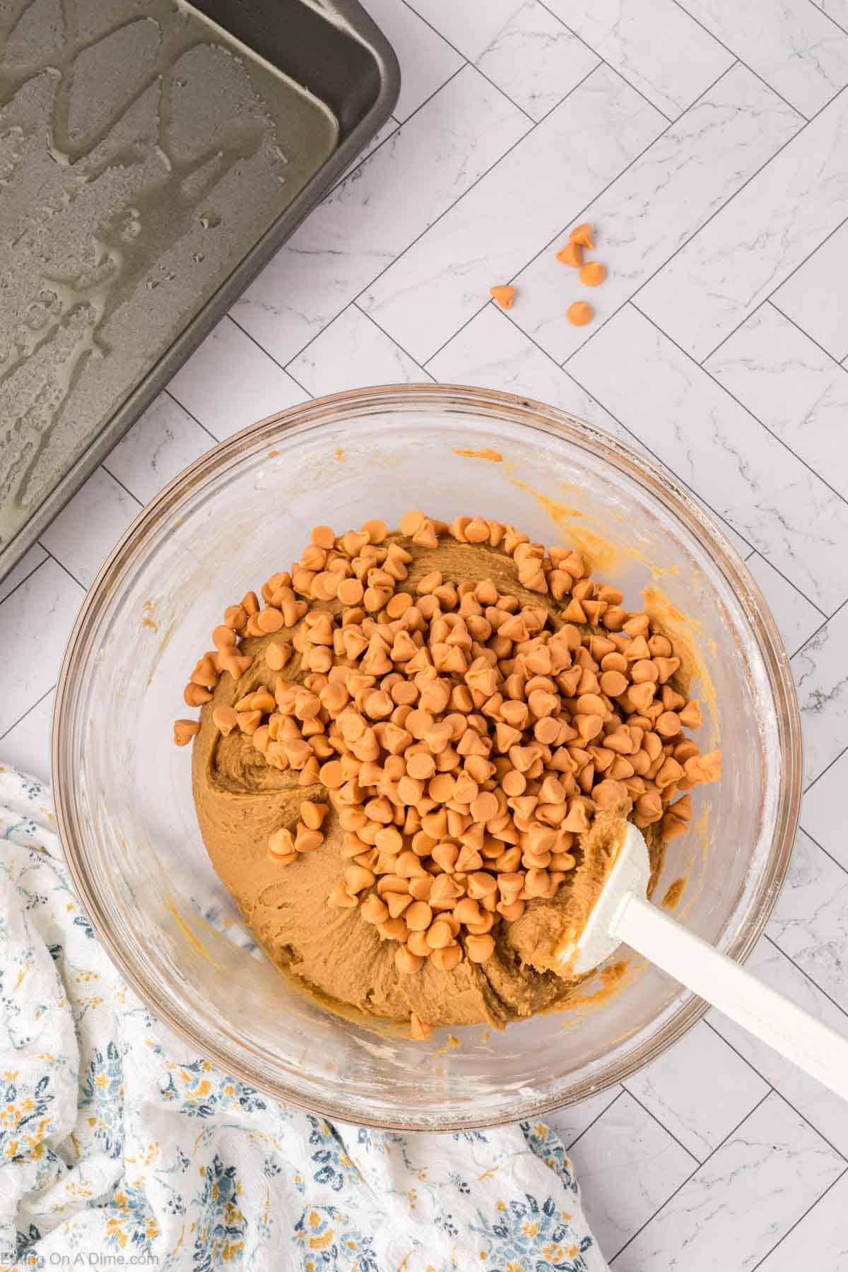 A glass bowl filled with cookie dough and butterscotch chips sits on a white, patterned countertop, ready to become delicious butterscotch brownies. A white spatula rests in the bowl, with a metal baking pan and floral cloth nearby.
