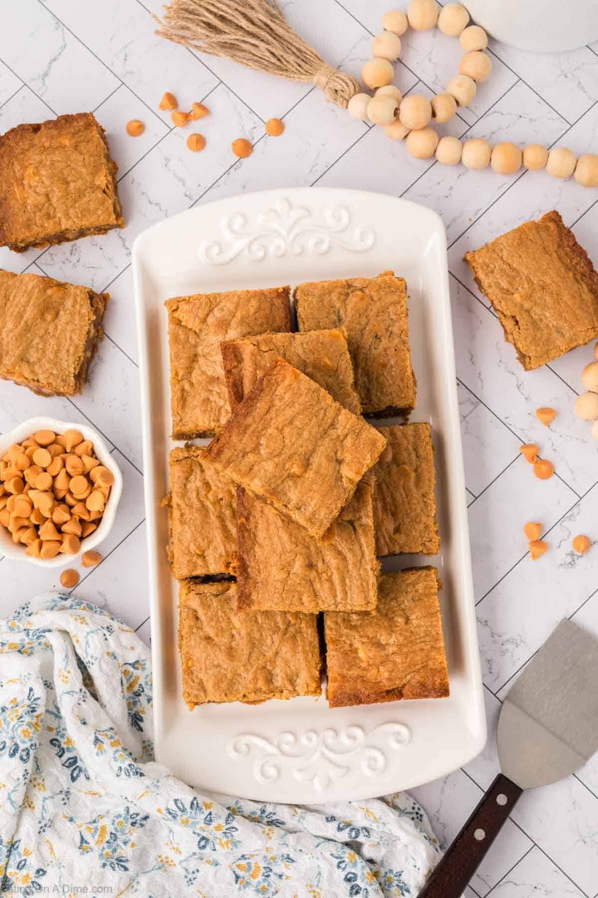 A rectangular white platter holds a stack of golden-brown butterscotch brownies. Butterscotch chips are scattered around, with a small bowl of chips and a floral cloth nearby, all on a white patterned surface.