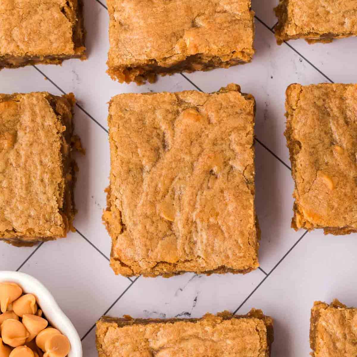 Top-down view of golden-brown butterscotch brownies with butterscotch chips, arranged on a white grid-patterned surface. A small bowl of butterscotch chips is visible in the corner.