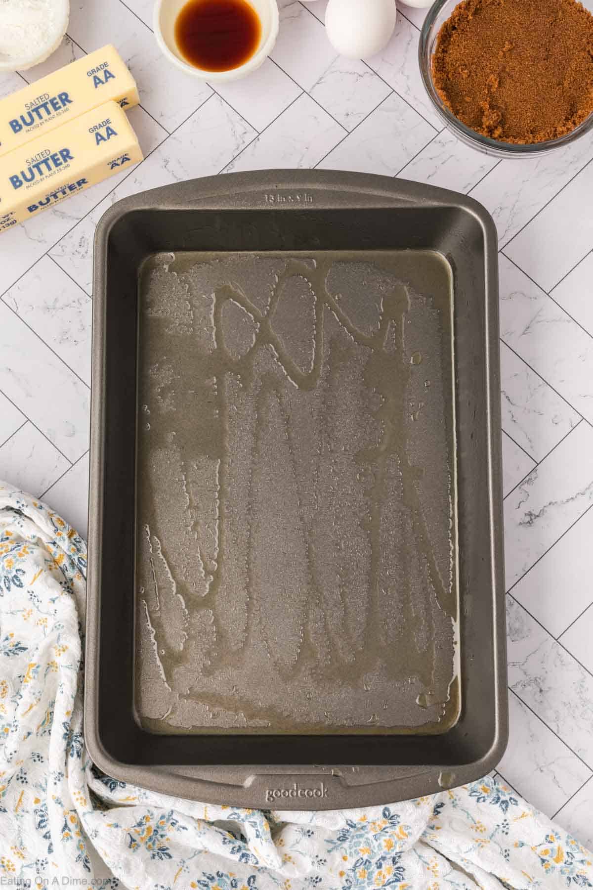 A greased rectangular baking pan sits on a marble countertop surrounded by ingredients for butterscotch brownies—sticks of butter, a bowl of brown sugar, vanilla extract, two eggs, and a floral-patterned cloth.