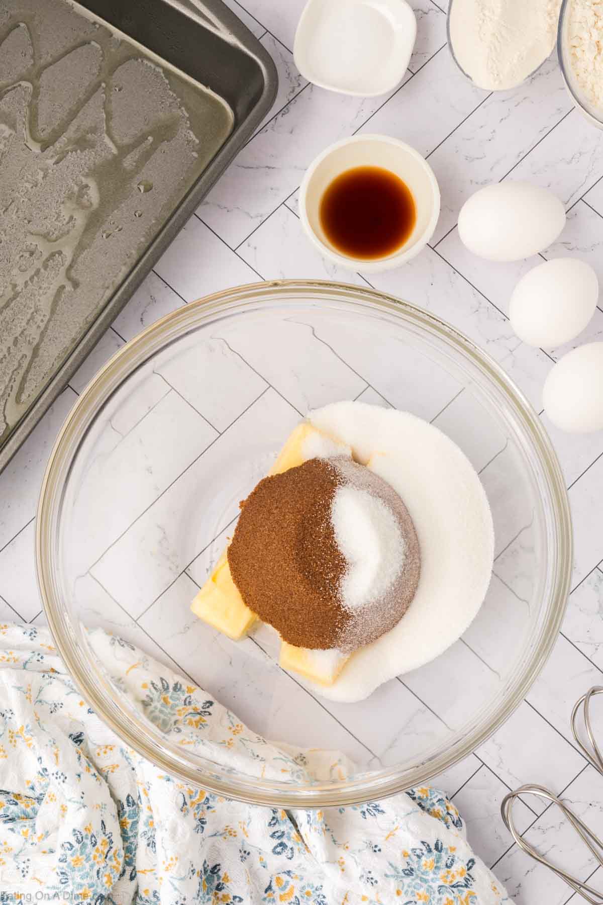 A glass mixing bowl with butter, brown sugar, and white sugar sits on a countertop—essentials for butterscotch brownies. Nearby are three eggs, vanilla extract, flour, a greased baking pan, and a floral kitchen towel.
