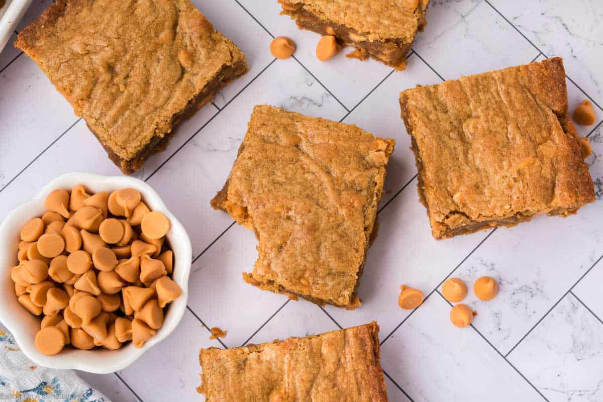 Five golden-brown butterscotch brownies are arranged on a tiled surface with a small white bowl of butterscotch chips and a few scattered chips nearby. The brownies look soft and chewy with a slightly cracked top.
