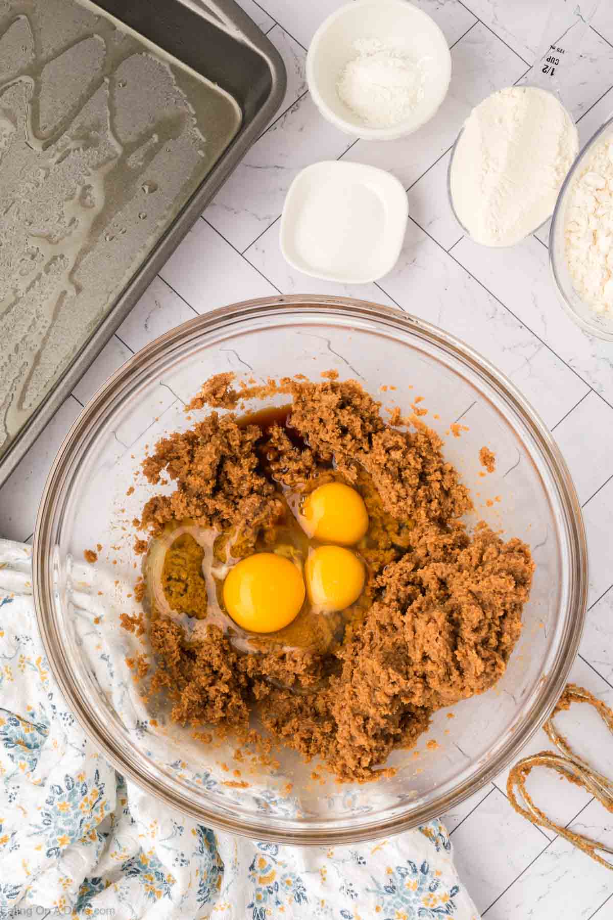 A glass bowl with brown sugar, three egg yolks, and vanilla extract sits on a countertop next to a greased baking pan and small bowls of flour and baking powder—ingredients perfect for making butterscotch brownies.