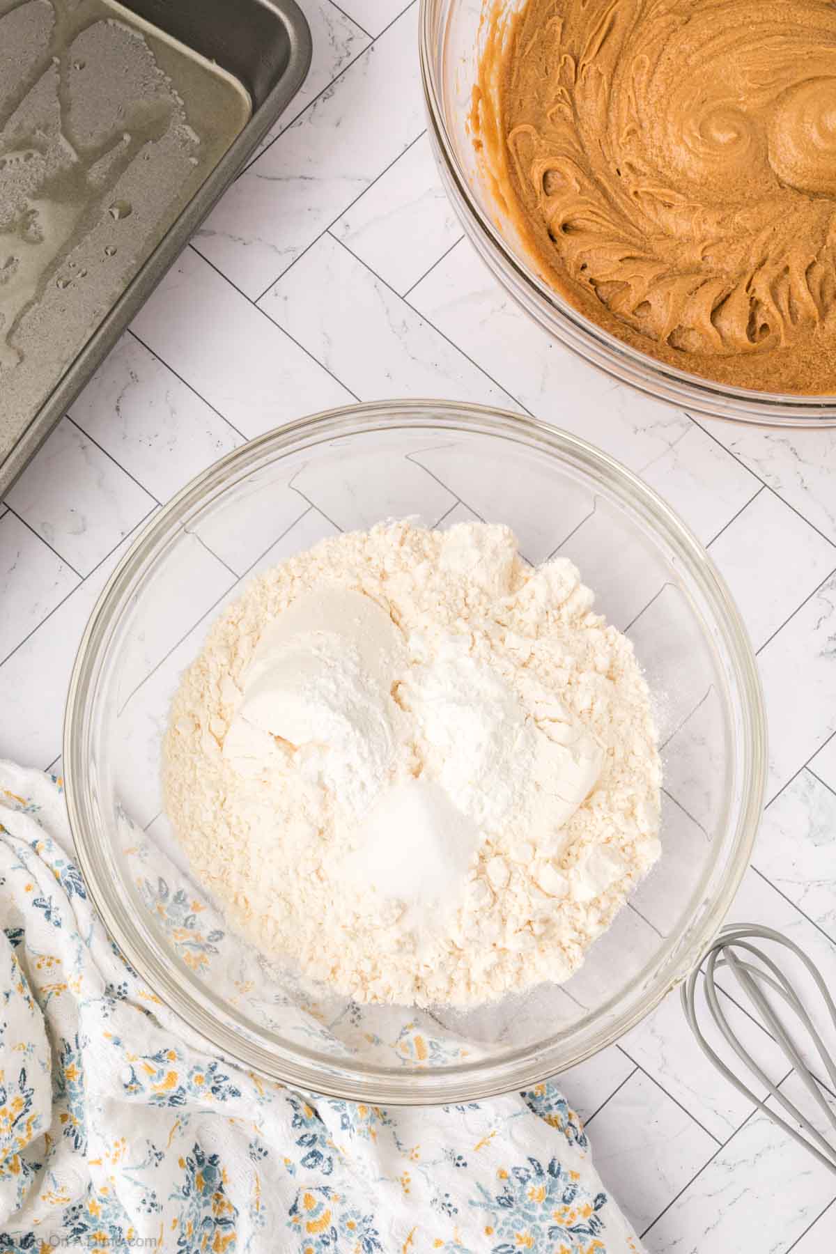A glass bowl with flour, baking powder, and baking soda sits on a white countertop next to a bowl of butterscotch brownies batter, a greased baking pan, a whisk, and a floral-patterned cloth.