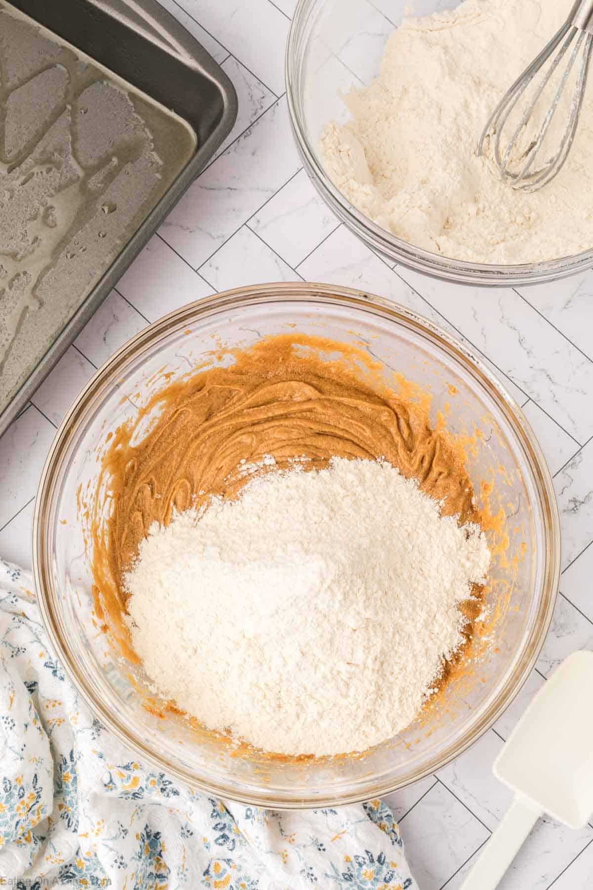 A glass bowl with butterscotch brownies cookie dough and flour being mixed, a whisk in a bowl of flour, a greased baking pan, and a floral cloth on a white tiled surface.