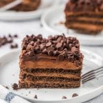 A square slice of Chocolate Icebox Cake with several layers, topped with chocolate chips, sits on a white plate with a fork beside it. Other slices of this delicious dessert are visible in the background.