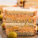 A stacked pimento cheese sandwich on white bread sits on a wooden board, held together with a toothpick. Green olives are placed around the pimento cheese sandwich for a classic Southern touch.