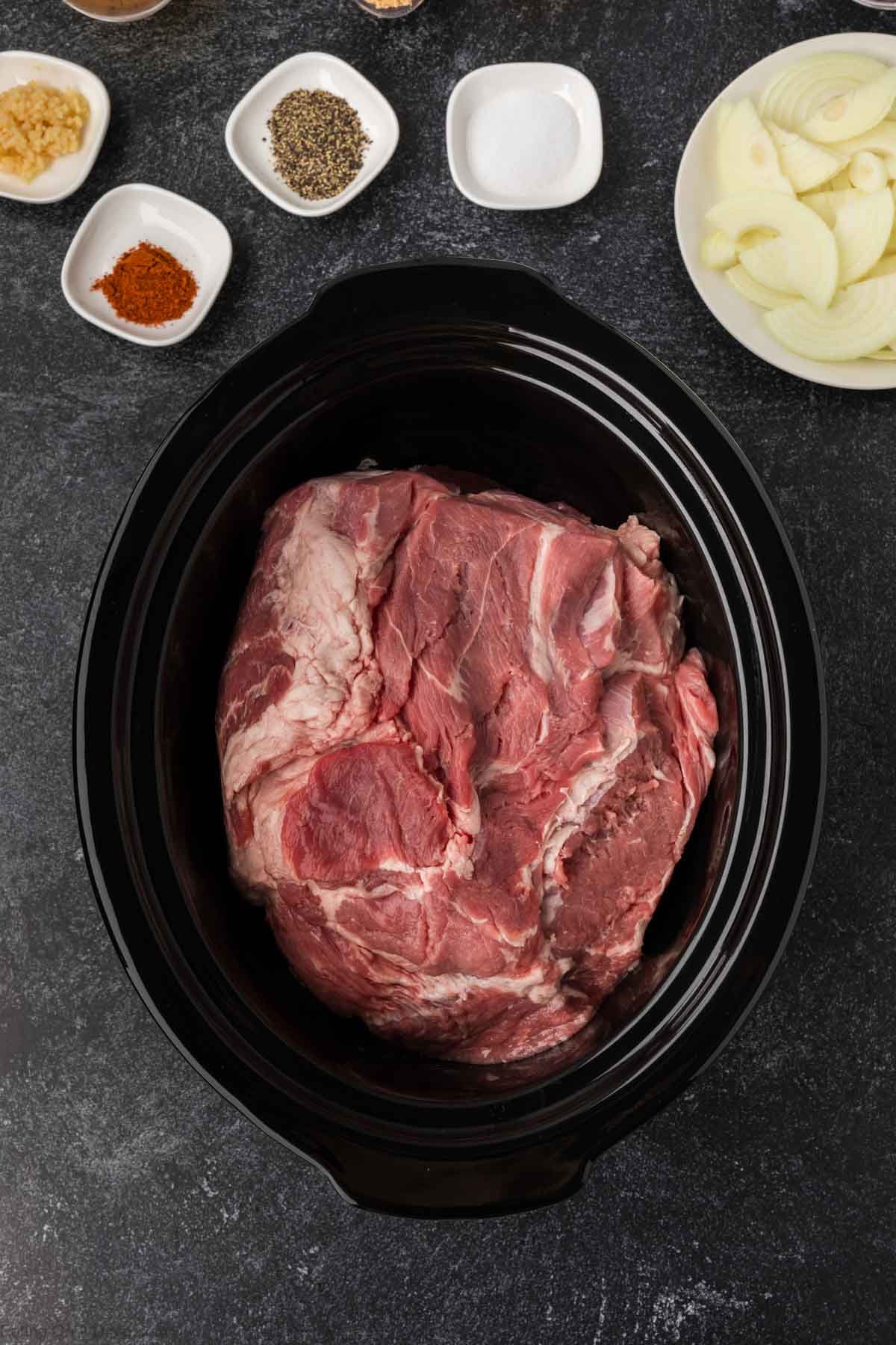 A large raw piece of beef sits in a black slow cooker, surrounded by small bowls of sliced onions, spices, salt, pepper, and minced garlic—perfect for transforming into a savory dish inspired by a Pulled Pork Crock Pot Recipe.