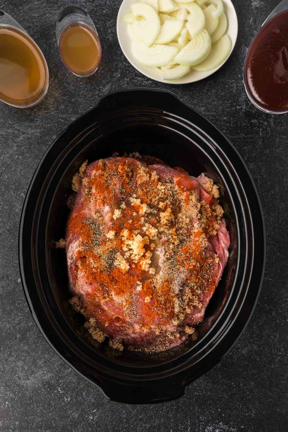 A seasoned raw pork roast sits in a black slow cooker, ready to become a delicious Pulled Pork Crock Pot Recipe, surrounded by bowls of sliced onions, brown liquid, and reddish barbecue sauce on a dark countertop.