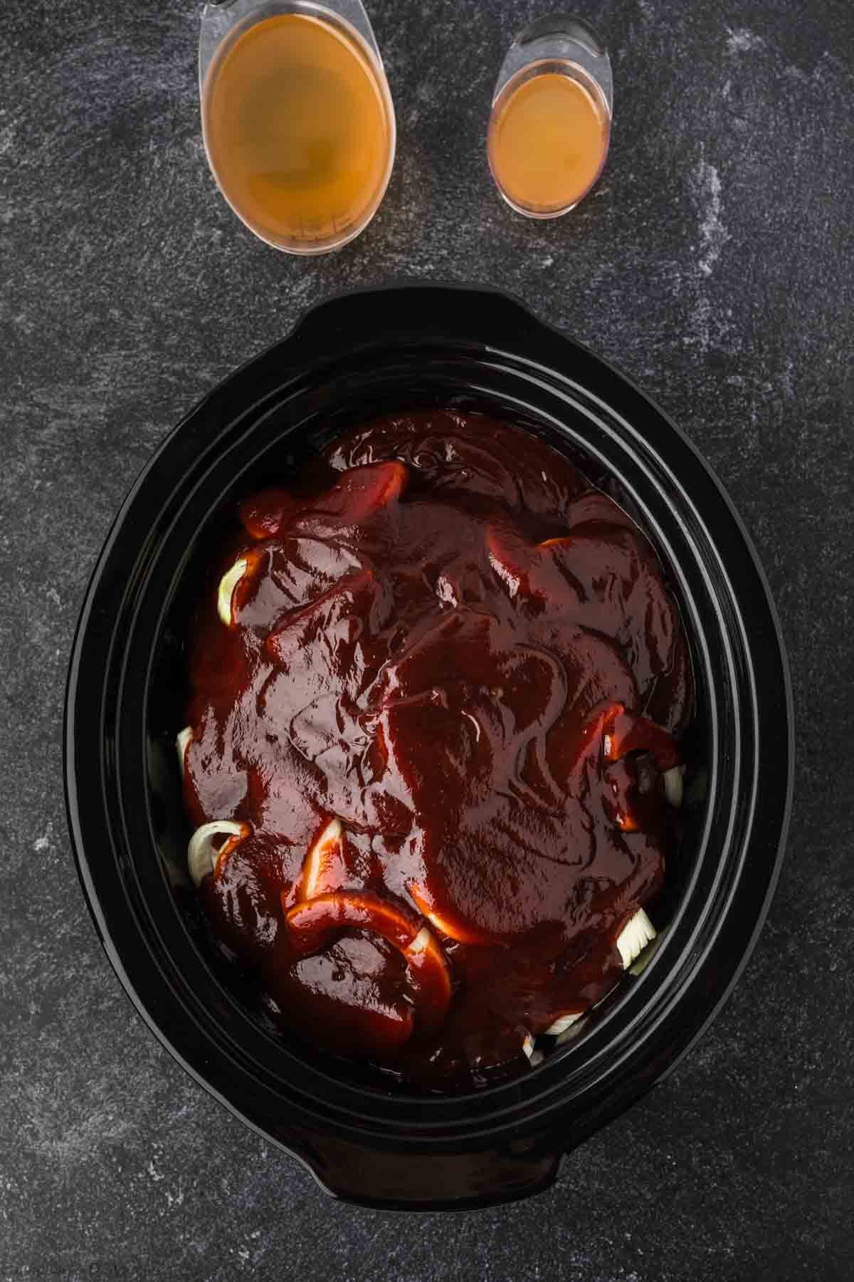 A slow cooker, perfect for a Pulled Pork Crock Pot Recipe, is filled with chicken, sliced onions, and barbecue sauce on a dark countertop. Two glasses filled with a light brown liquid sit above the slow cooker.